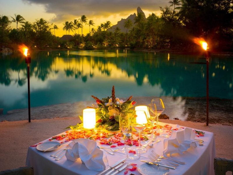 A beautifully set dinner table adorned with flowers, candles, and a view of a tranquil lagoon at sunset.