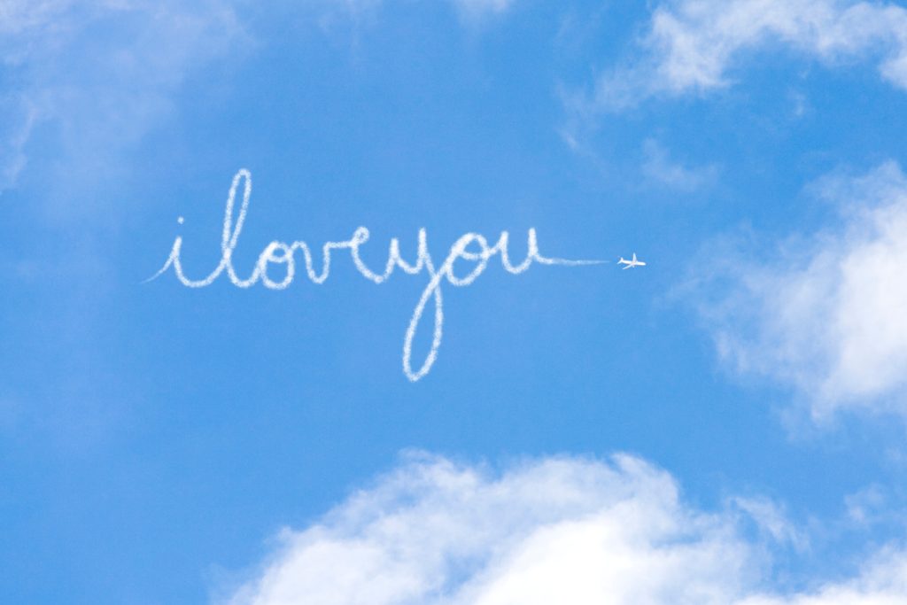 Skywriting forms the phrase "I love you" against a blue sky with fluffy clouds, symbolizing romance and affection.
