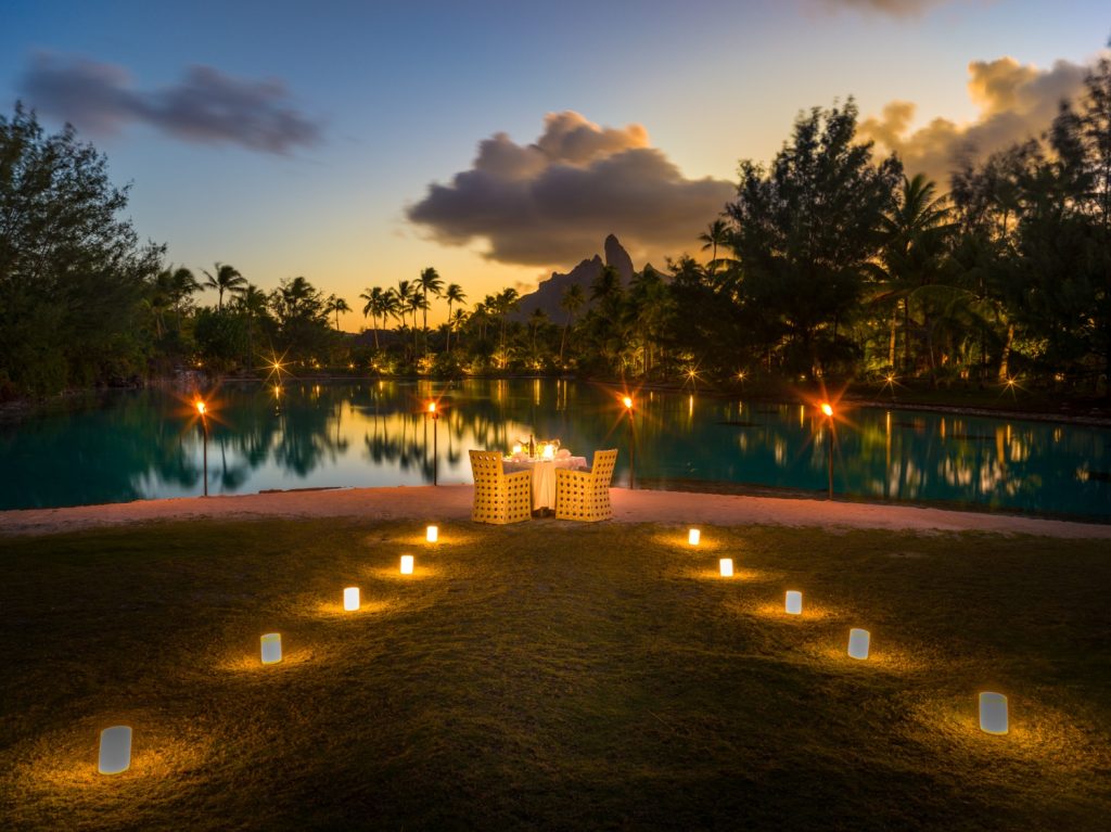 A romantic dining setup by a tranquil pond, illuminated by lanterns at sunset surrounded by lush tropical foliage.