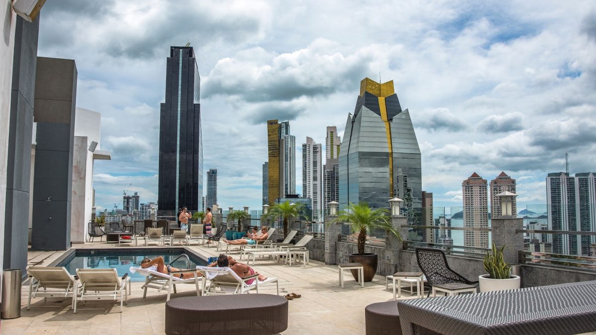 Rooftop pool area with lounging guests, surrounded by modern skyscrapers and a cloudy sky in a vibrant urban setting.