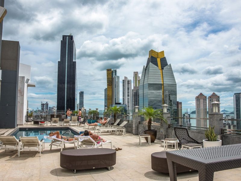 Rooftop pool area with lounging guests, surrounded by modern skyscrapers and a cloudy sky in a vibrant urban setting.