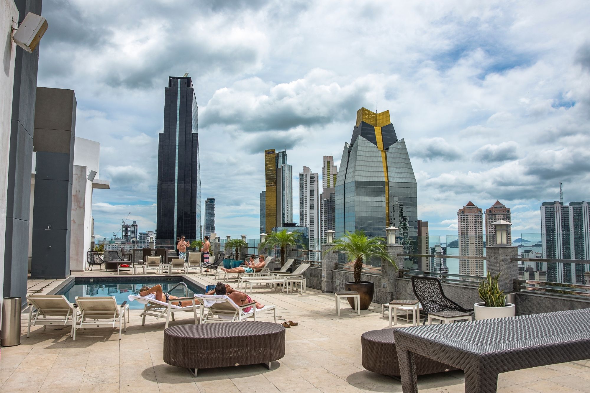 Rooftop pool area with lounging guests, surrounded by modern skyscrapers and a cloudy sky in a vibrant urban setting.