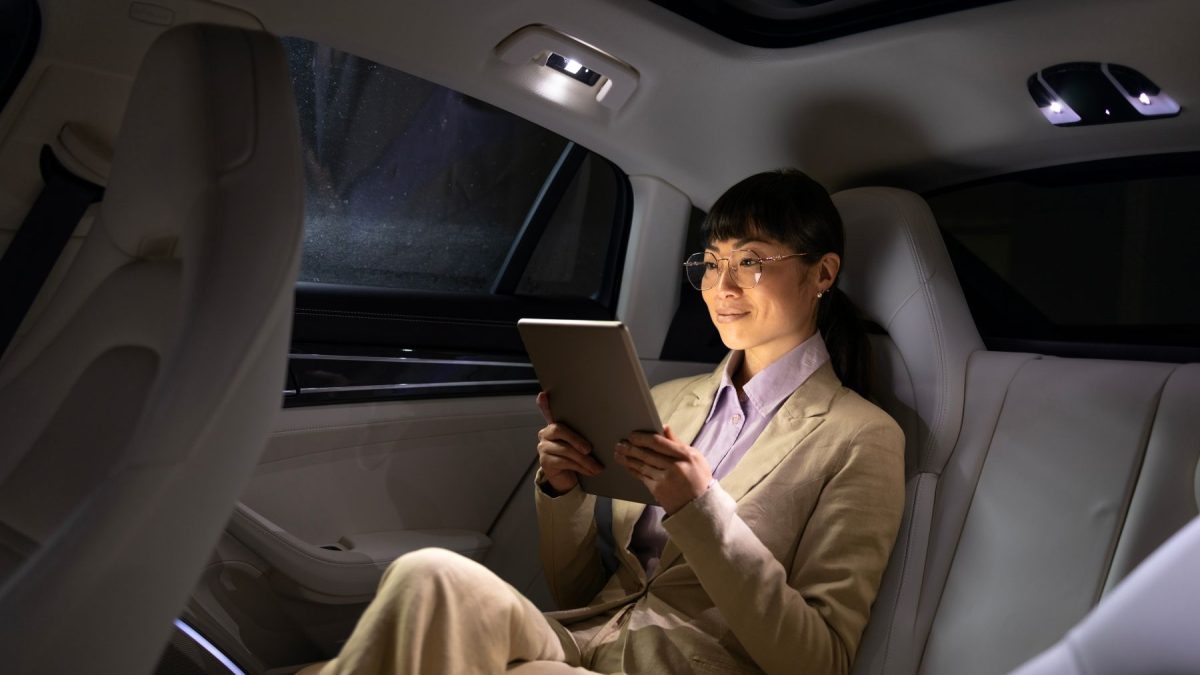 A woman in a beige suit relaxes in the backseat of a luxury vehicle, reading on a tablet.