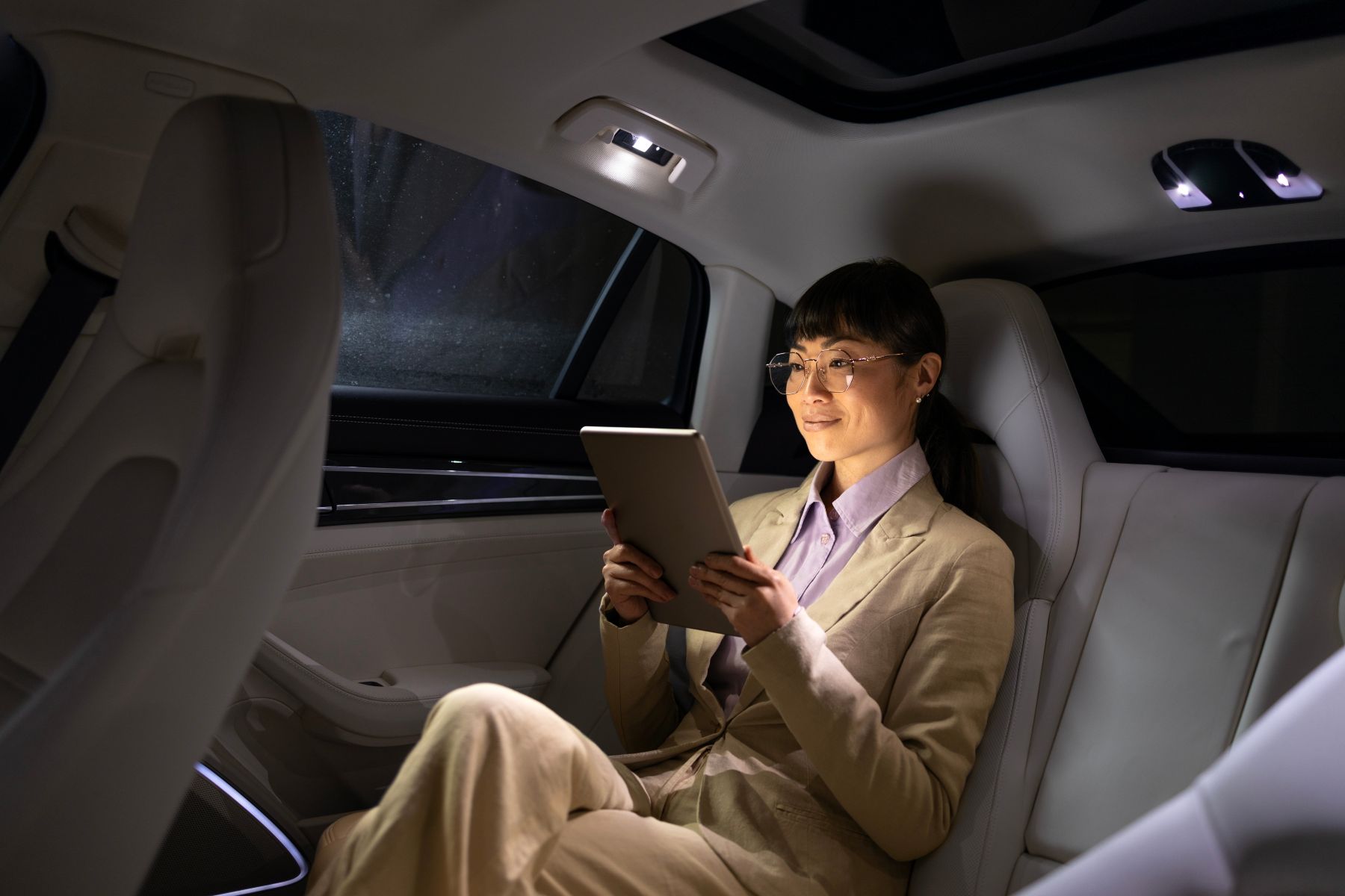 A woman in a beige suit relaxes in the backseat of a luxury vehicle, reading on a tablet.