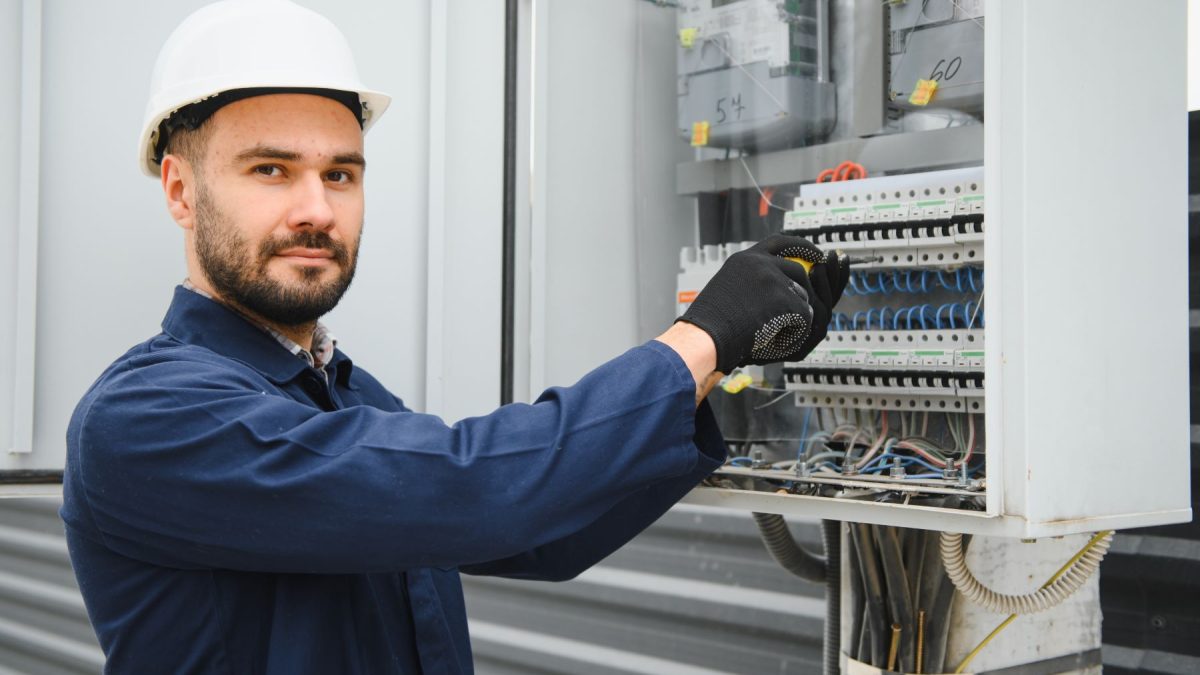Electrician working on a control panel, wearing a hard hat and gloves, inspecting electrical connections and components.