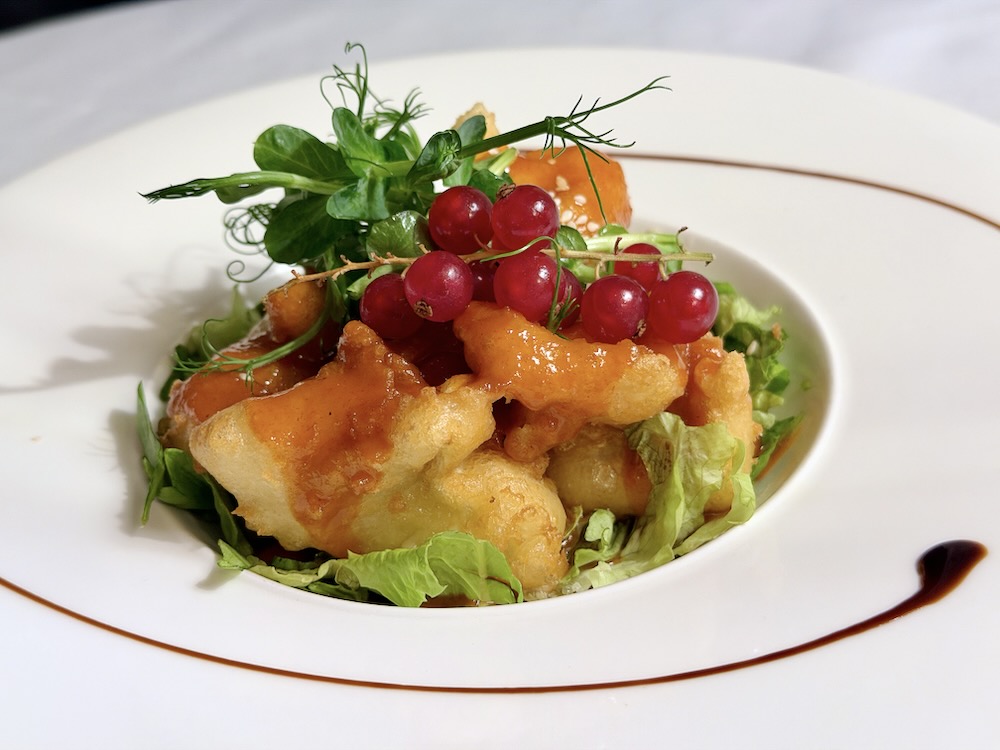 A plate of lightly battered and fried delicacies topped with a sweet sauce, garnished with red currants and fresh greens.