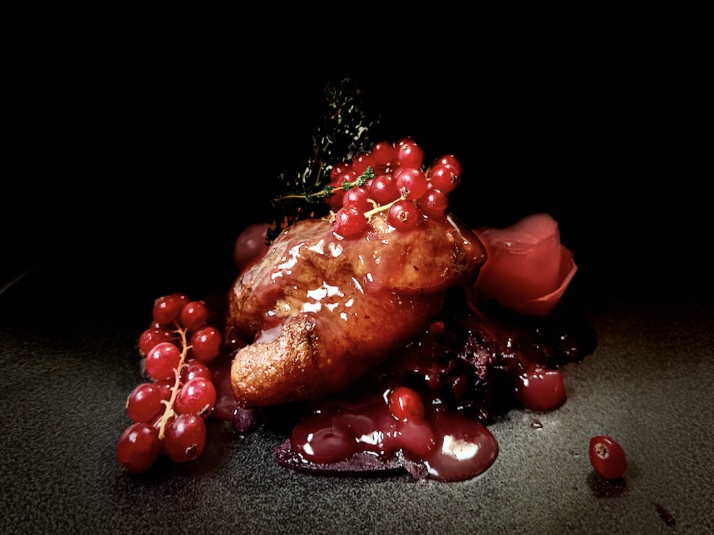 A gourmet dish featuring glazed meat topped with fresh berries and herbs against a dark background.