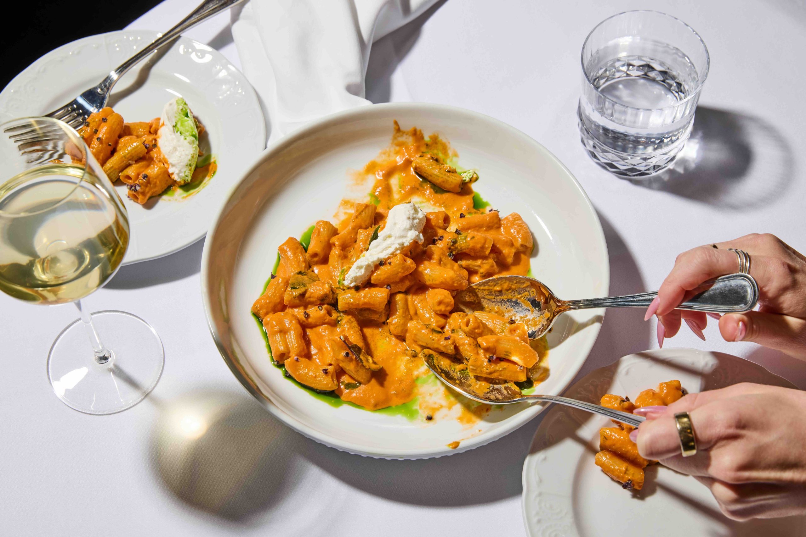 Spicy rigatoni garnished with fresh ricotta, served with a glass of white wine on a table set for dinner.