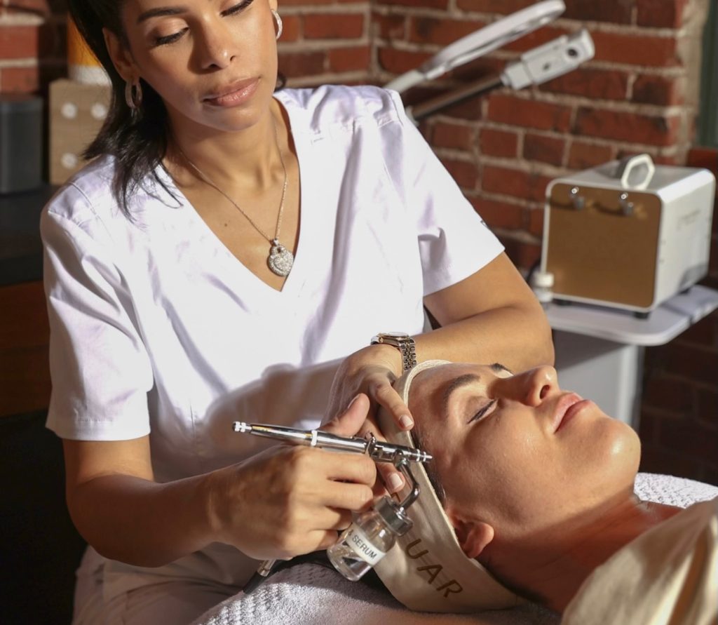 A skin care professional uses an airbrush to apply serum to a client's face in a spa setting.