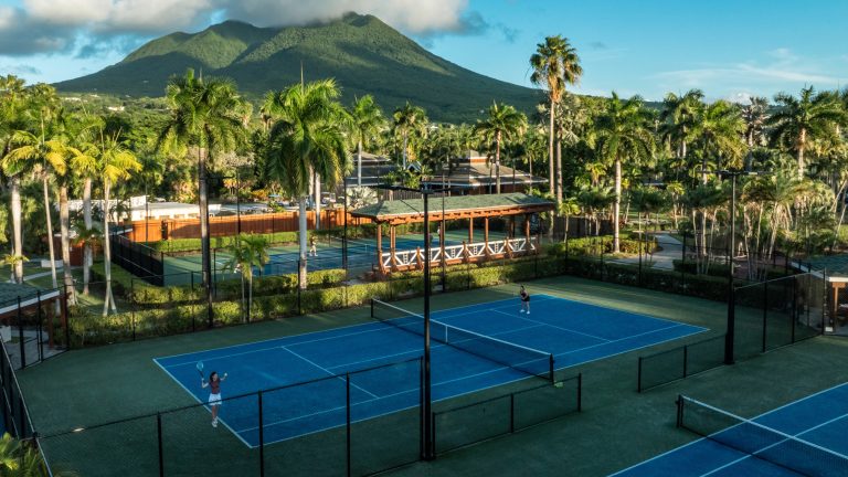 Children participate in a tennis camp at Four Seasons Resort Nevis, with lush palm trees and mountains in the background.