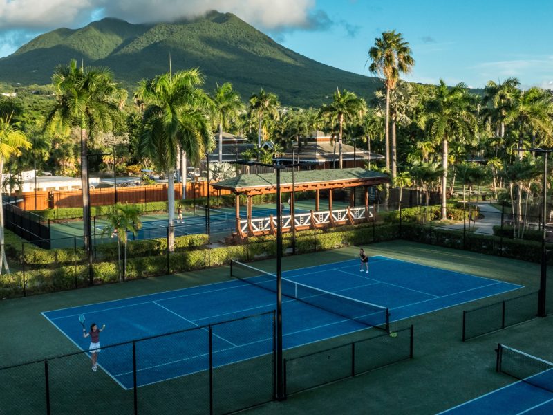 Children participate in a tennis camp at Four Seasons Resort Nevis, with lush palm trees and mountains in the background.