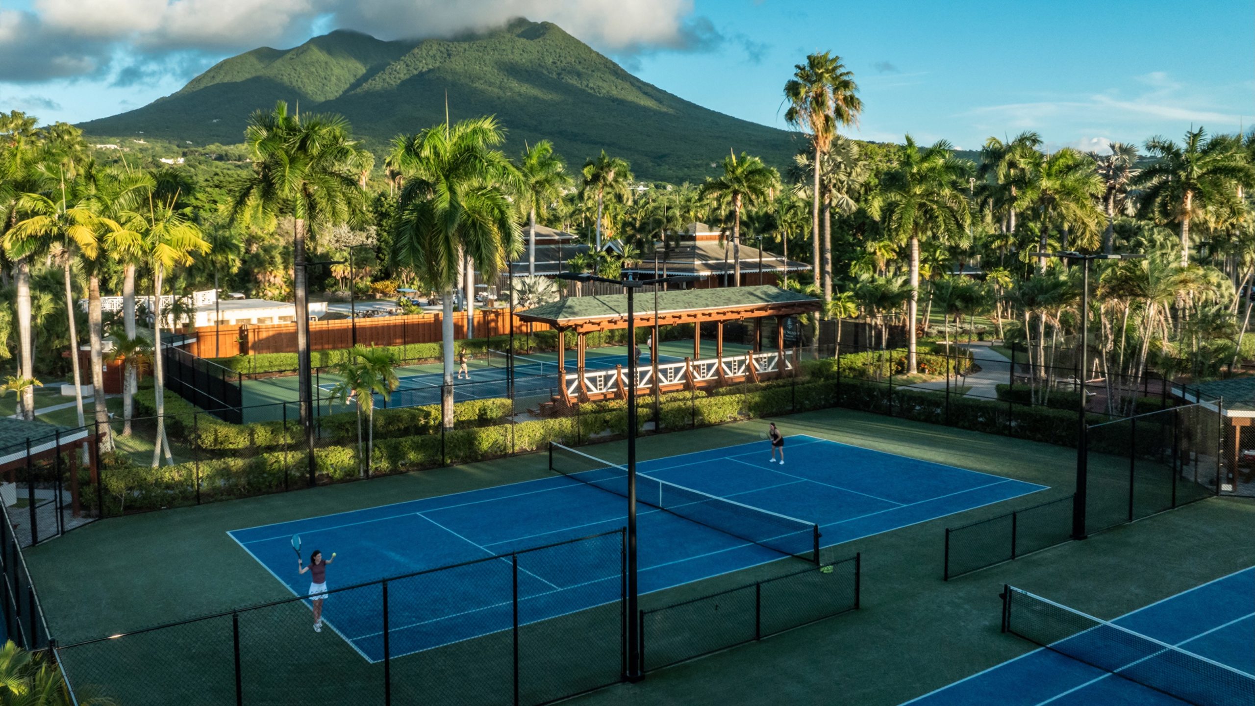 Children participate in a tennis camp at Four Seasons Resort Nevis, with lush palm trees and mountains in the background.