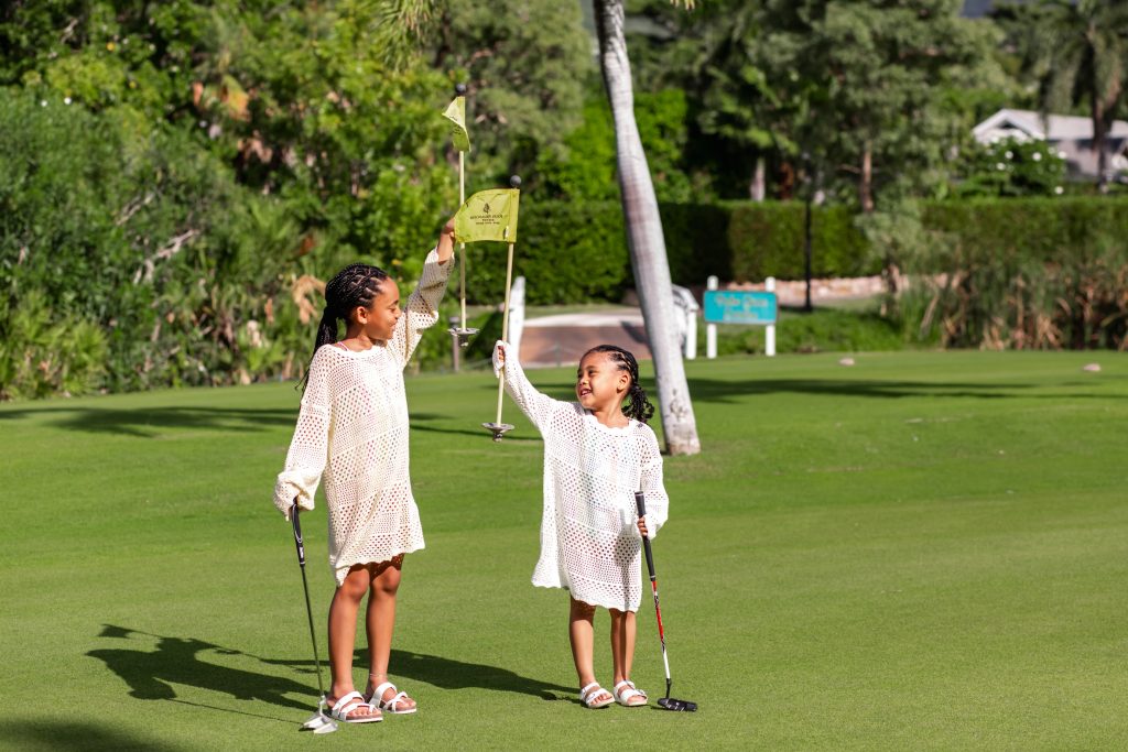 Two girls in matching outfits hold up golf flags on a sunny green, smiling and enjoying their time on the course.