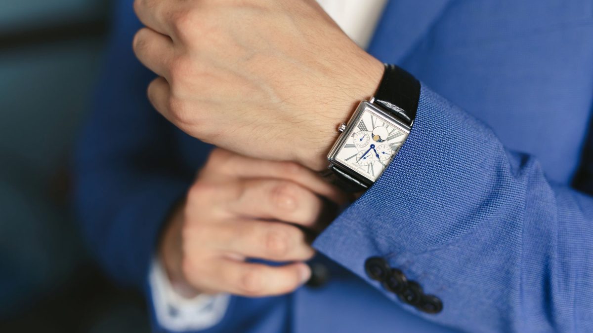 A close-up of a hand adjusting a stylish watch on the wrist, paired with a blue suit jacket.