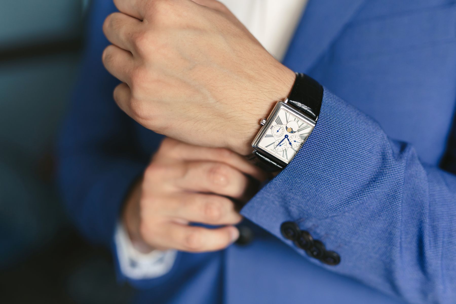 A close-up of a hand adjusting a stylish watch on the wrist, paired with a blue suit jacket.