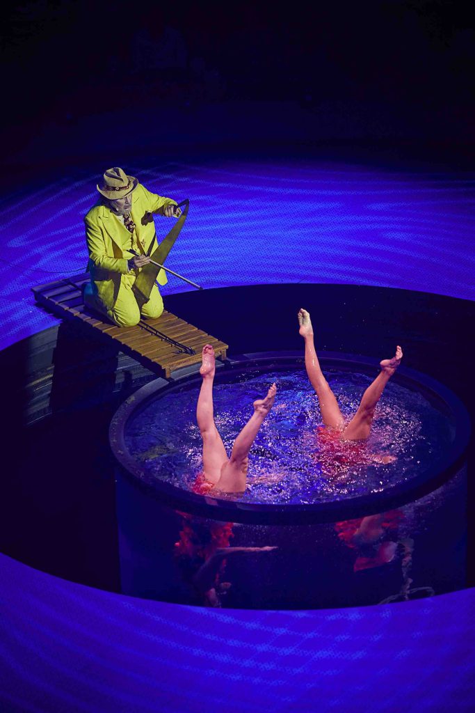 A performer in a yellow outfit plays a musical saw while two acrobats with legs raised immerse in a circular pool.