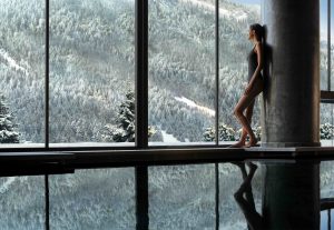 Woman in swimwear relaxing by a large window with a snowy mountain view, reflecting in the water below.