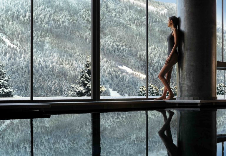 Woman in swimwear relaxing by a large window with a snowy mountain view, reflecting in the water below.