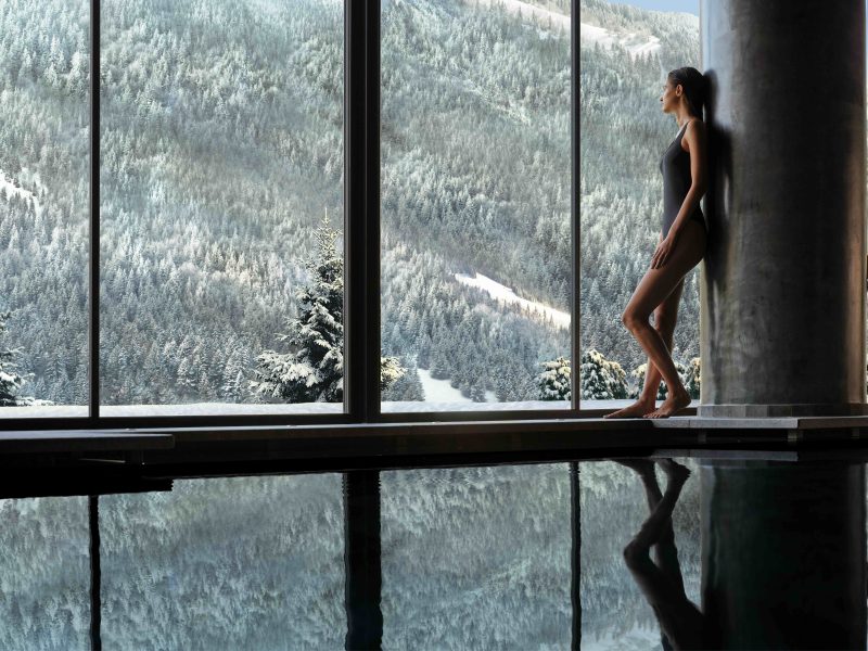 Woman in swimwear relaxing by a large window with a snowy mountain view, reflecting in the water below.