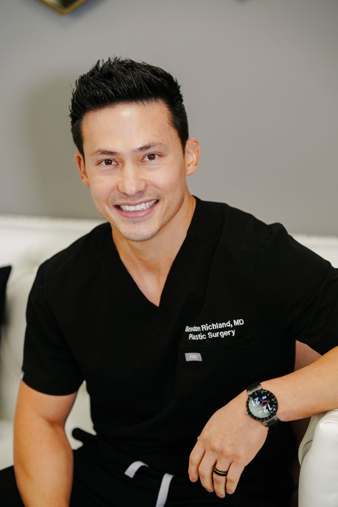 Smiling doctor in scrubs, looking confidently at the camera, seated on a couch in a modern medical office.
