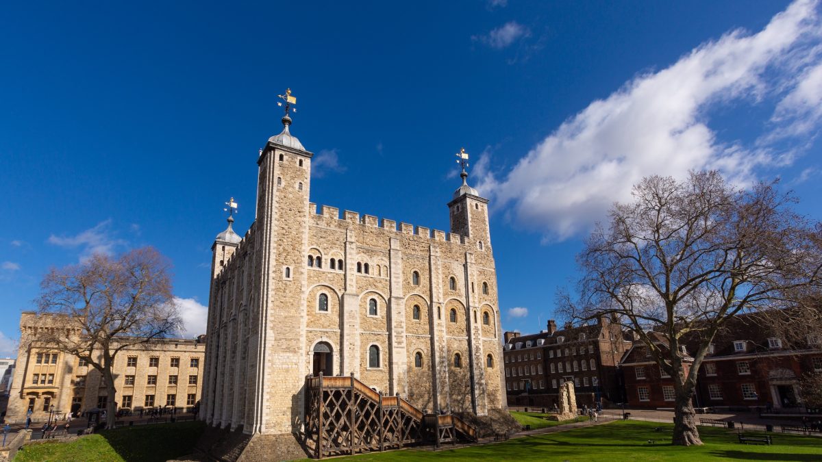 The Tower of London stands majestically under a clear blue sky, surrounded by lush green lawns and historic buildings.