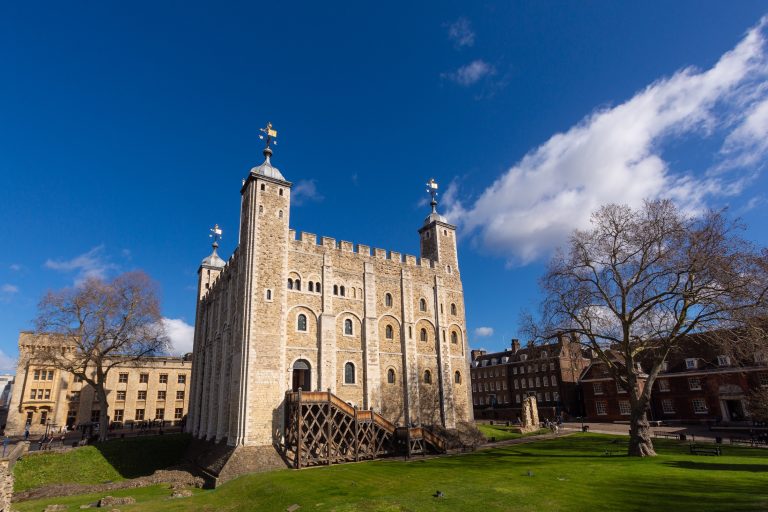 The Tower of London stands majestically under a clear blue sky, surrounded by lush green lawns and historic buildings.