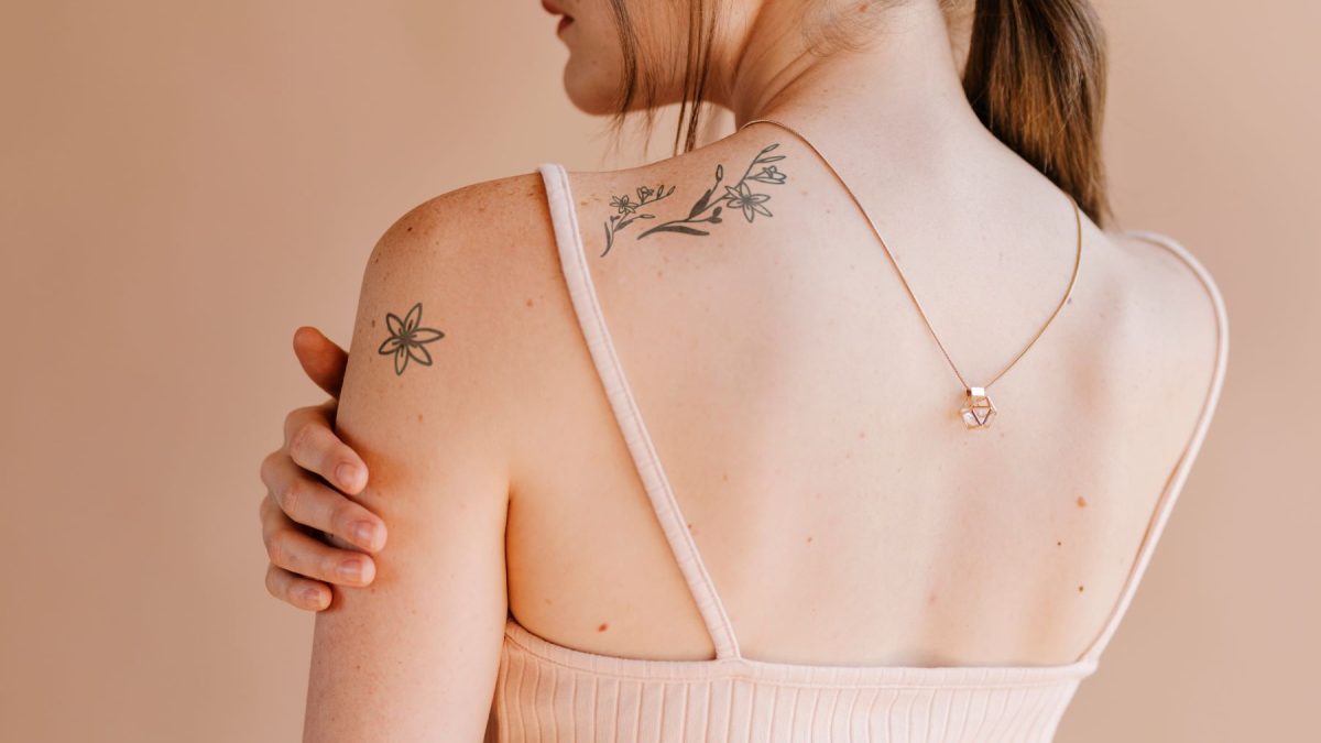 A woman with floral tattoos on her shoulder and back, wearing a light tank top, showcasing a minimalist necklace.