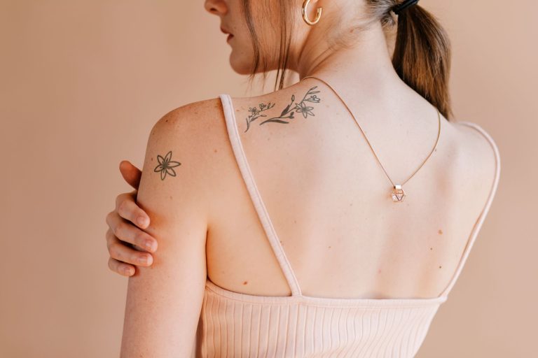 A woman with floral tattoos on her shoulder and back, wearing a light tank top, showcasing a minimalist necklace.