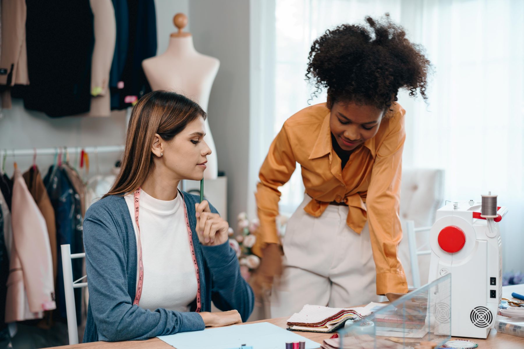 Two women collaborate in a clothing design studio, discussing fabrics and quality while a sewing machine is visible nearby.