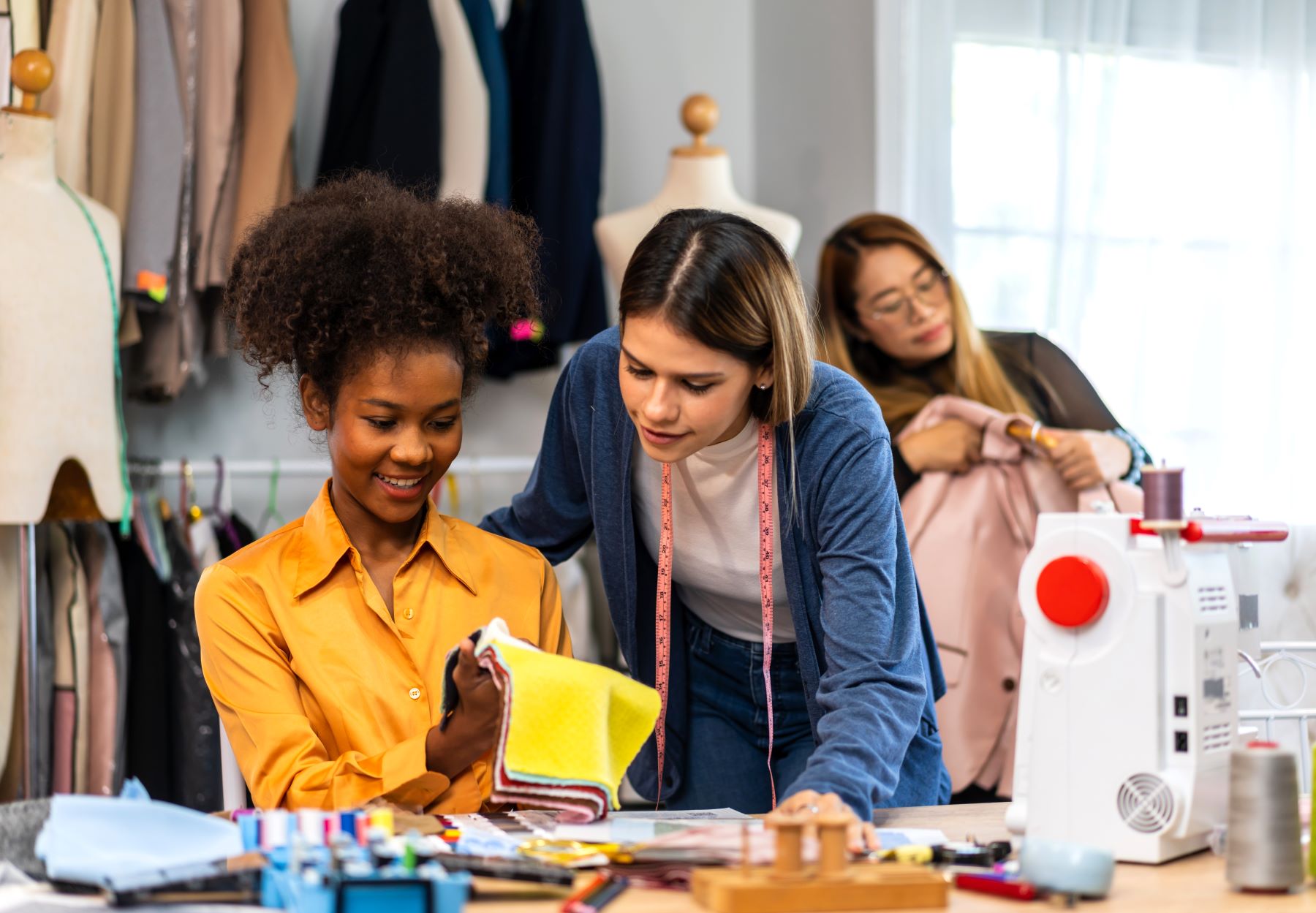 Two women examine fabric samples in a clothing workshop, while another woman adjusts a garment in the background.