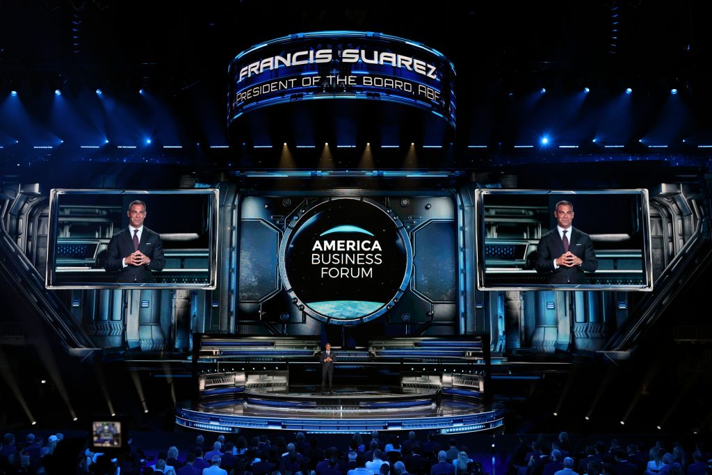 Francis Suarez speaks at the America Business Forum on a high-tech stage, with an audience visible in the foreground.
