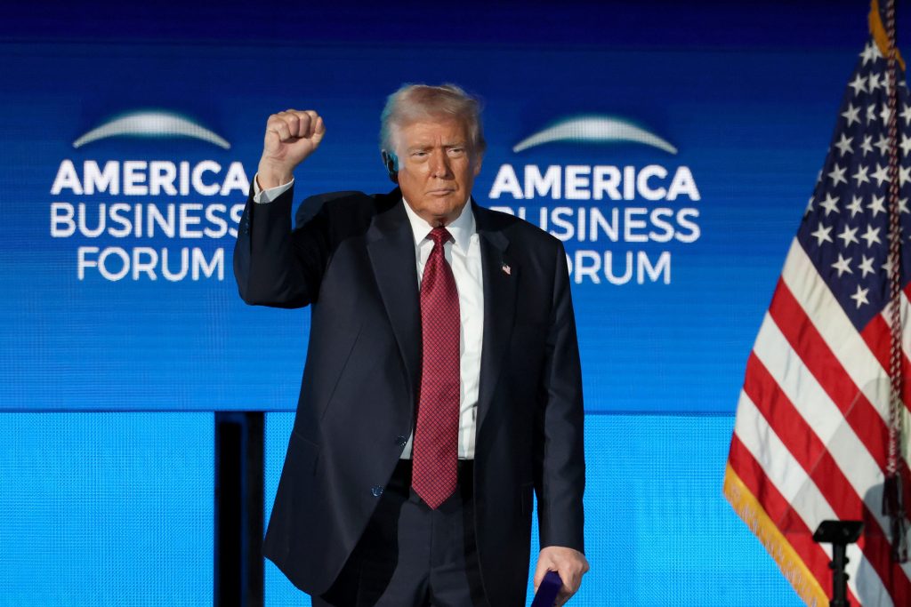 A speaker raises a fist at the America Business Forum, with an American flag in the background.