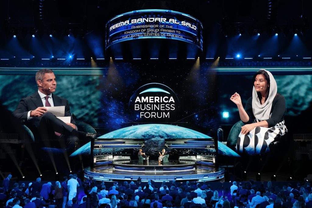A speaker discussion at the America Business Forum features two presenters on a futuristic stage with an engaged audience.