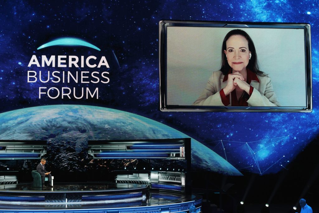 A virtual speaker participates in the America Business Forum, with a host seated in the studio below.