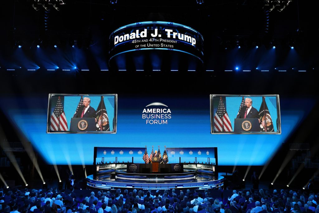 Donald Trump speaks at the America Business Forum, with flags and a large screen displaying his image in a vibrant venue.