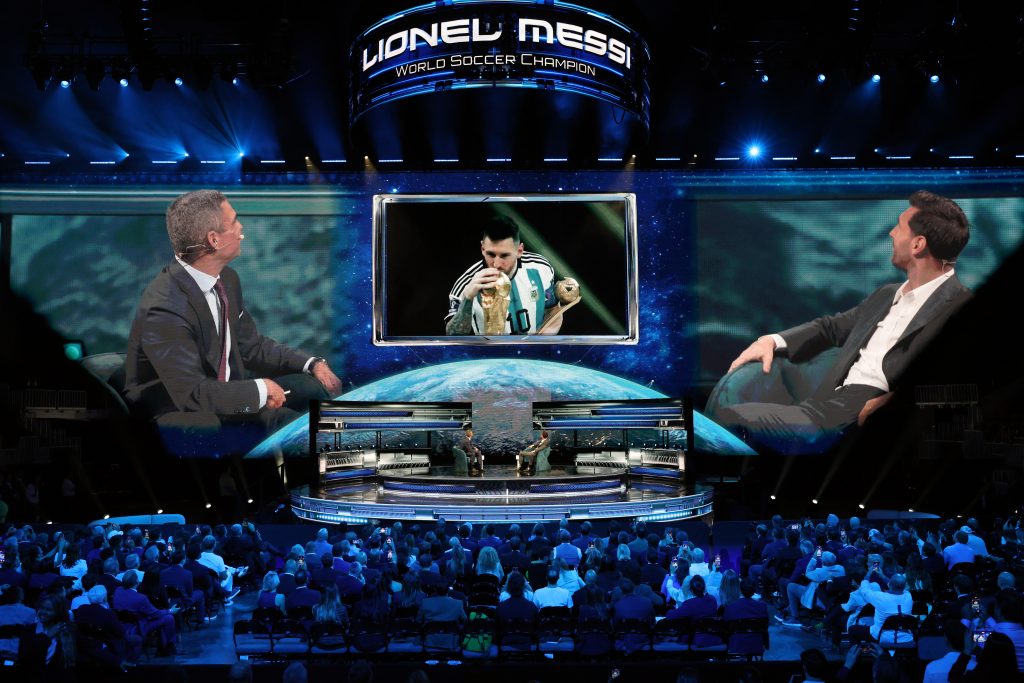 Lionel Messi is featured on a large screen during the America Business Forum in Miami, alongside two seated hosts.