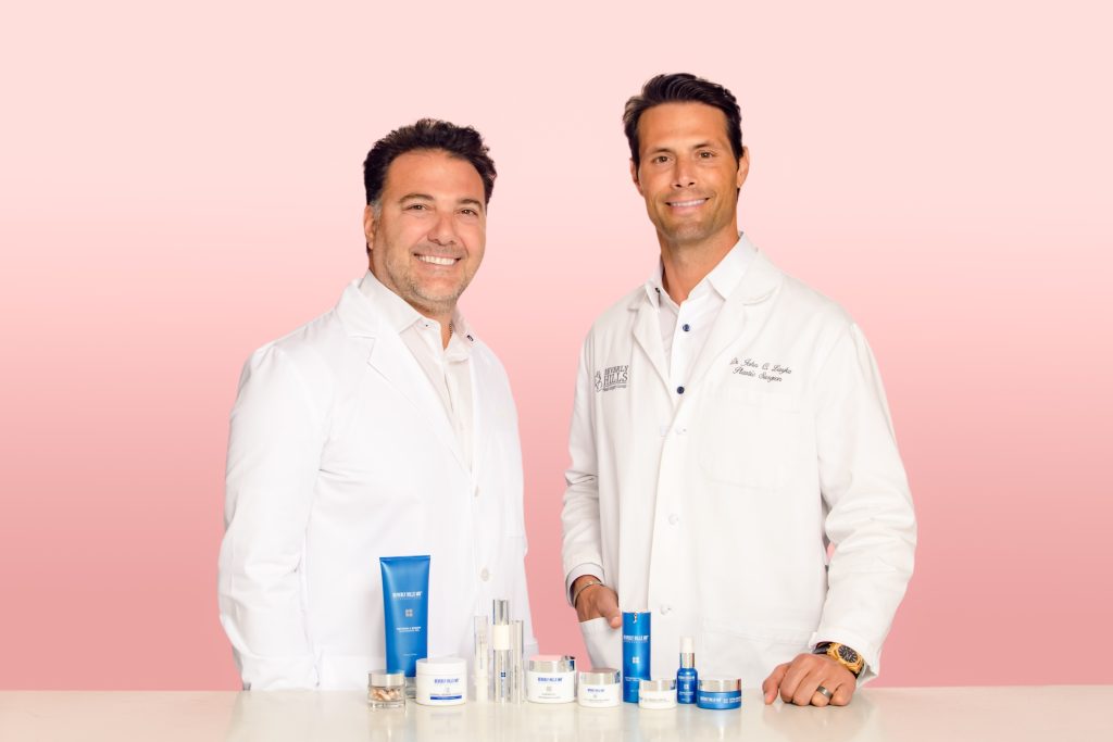 Two men in white lab coats pose with Beverly Hills MD skincare products against a pink background.