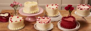 A display of heart-shaped cakes adorned with romantic decorations, surrounded by pink roses on a rustic wooden table.