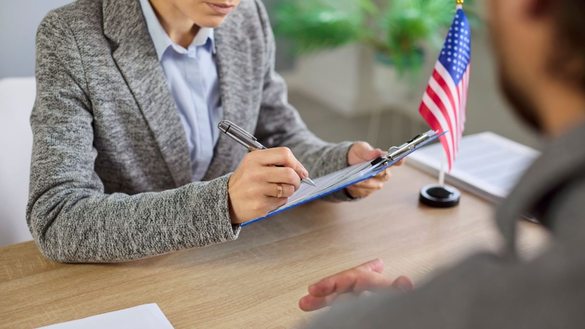 A professional discussing visa application details with a client at a desk, with a small American flag in the background.