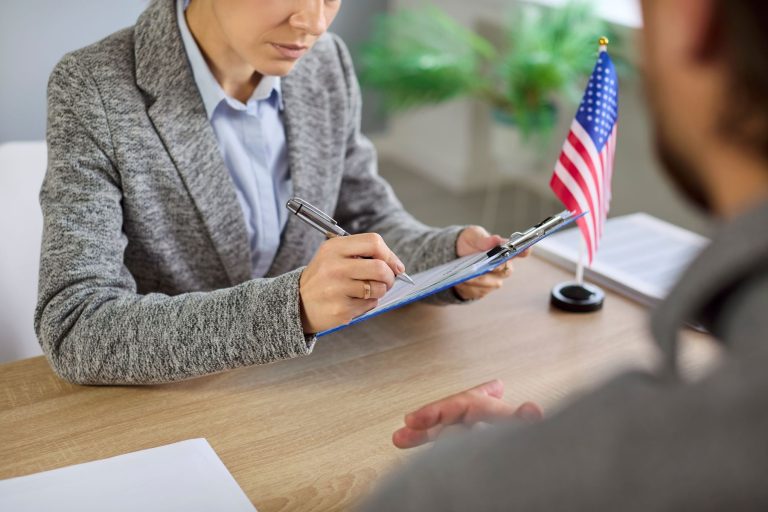 A professional discussing visa application details with a client at a desk, with a small American flag in the background.