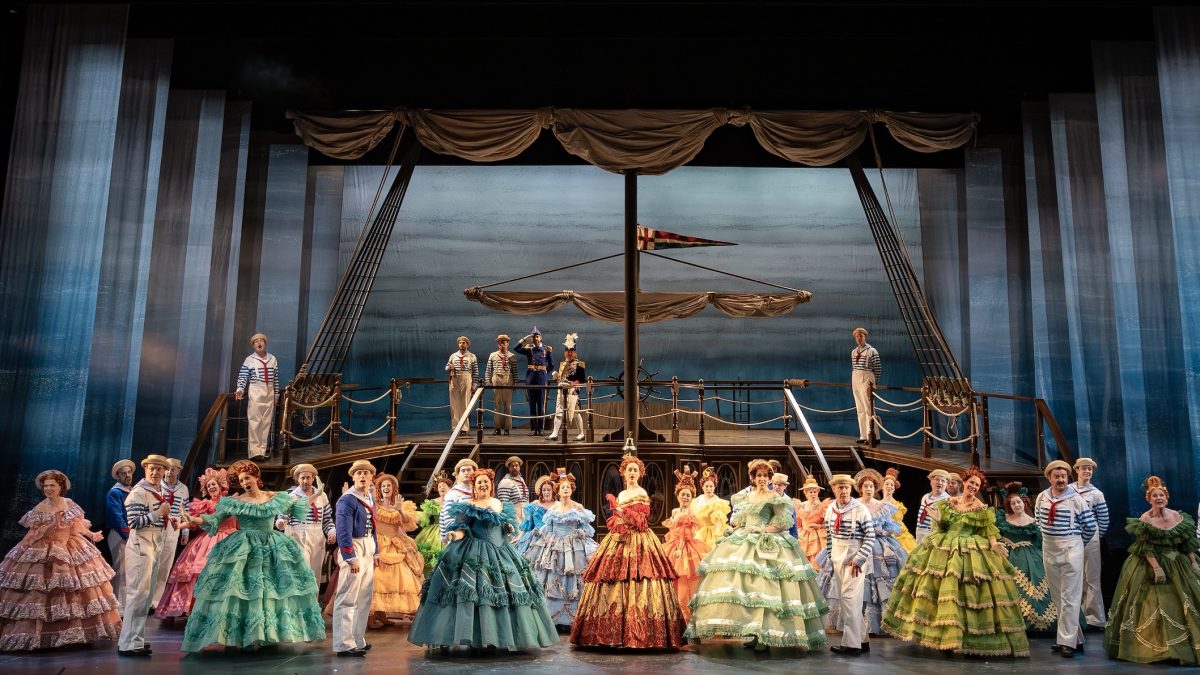 The cast of ENO's HMS Pinafore performs in colorful costumes on a ship stage, capturing a lively maritime atmosphere.