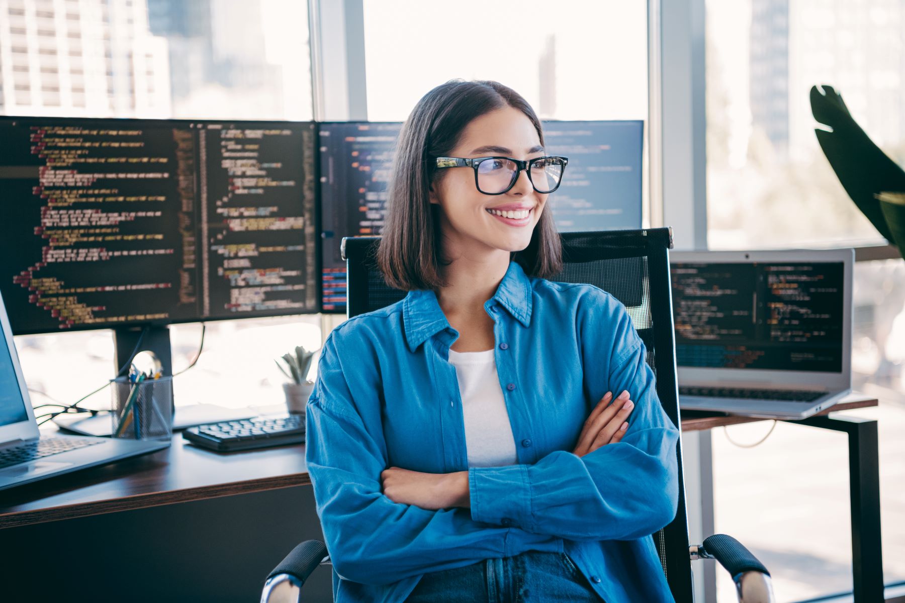 Woman in a blue shirt smiling confidently in an office setting with multiple computer screens displaying code.