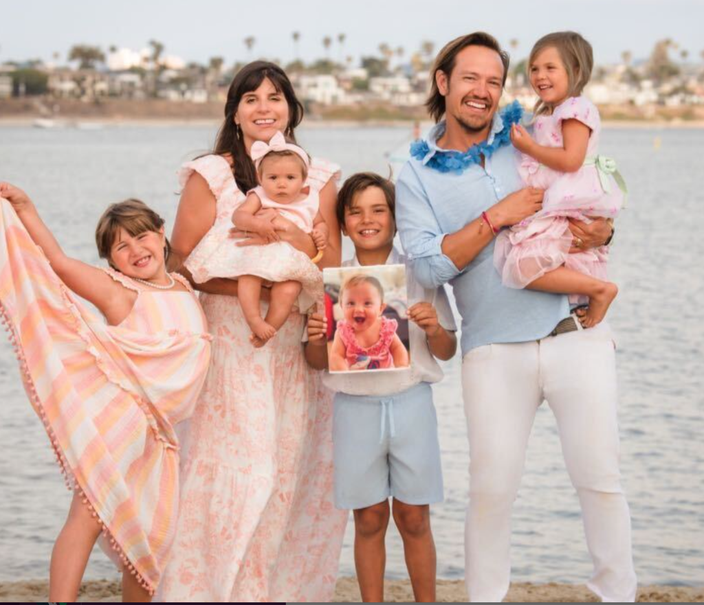 A family with three children poses by the water, two of them holding a photograph of a smiling baby girl, celebrating parenting.