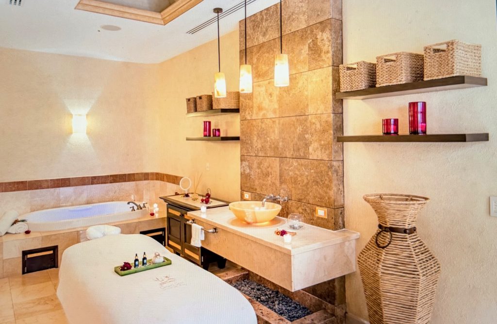 Luxurious spa room with a massage bed, soaking tub, and shelves adorned with candles and decorative baskets.