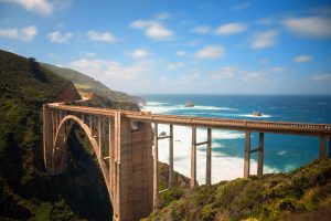A concrete arch bridge spans a lush, green coastal landscape with waves crashing against the shore below.
