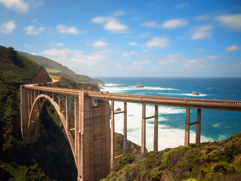 A concrete arch bridge spans a lush, green coastal landscape with waves crashing against the shore below.