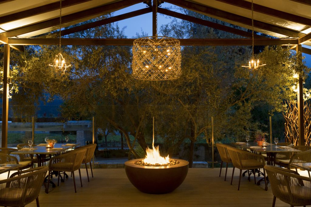 Cozy outdoor dining area with a fire pit, illuminated by stylish pendant lights and surrounded by greenery.