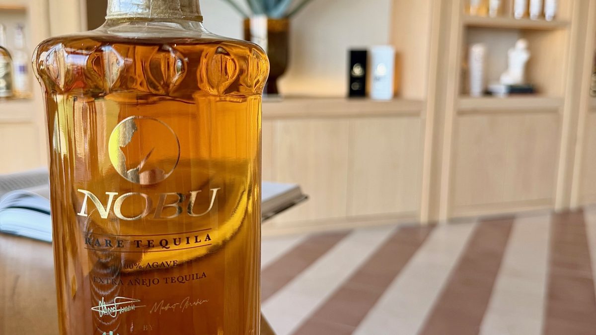 A bottle of Nobu rare añejo tequila on a wooden table, with a plant and shelves of tequila in the background.