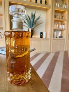 A bottle of Nobu rare añejo tequila on a wooden table, with a plant and shelves of tequila in the background.