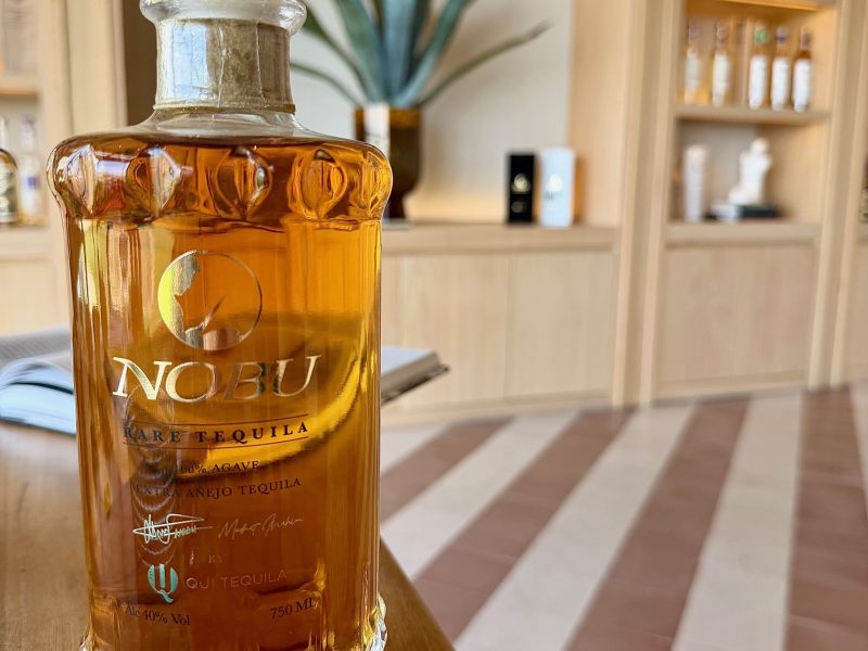 A bottle of Nobu rare añejo tequila on a wooden table, with a plant and shelves of tequila in the background.
